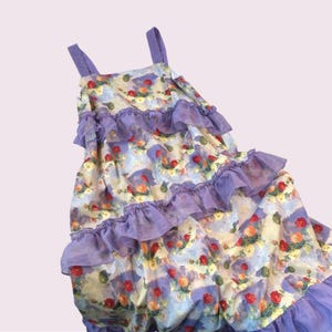 May include: A sleeveless dress with a floral pattern in shades of red, yellow, and green on a light blue and cream background. The dress features layers of purple ruffles and straps.