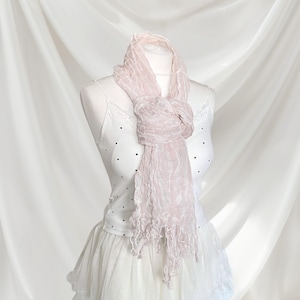 May include: A light pink, crinkled texture scarf with frayed ends draped around the neck of a white camisole featuring small black polka dots and lace trim, paired with a white skirt, displayed on a mannequin against a draped fabric backdrop.