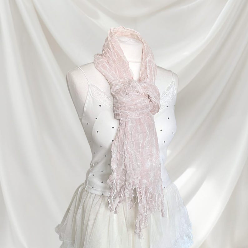 May include: A light pink, crinkled texture scarf with frayed ends draped around the neck of a white camisole featuring small black polka dots and lace trim, paired with a white skirt, displayed on a mannequin against a draped fabric backdrop.