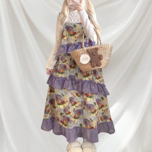 May include: A floral print dress with layered ruffles in lavender and a matching trim. The dress features a light yellow background with a colorful floral pattern. A woven tote bag with a teddy bear accent is also visible.