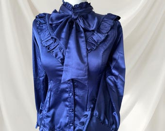 May include: A dark blue satin blouse featuring a high ruffled collar, a large bow tie at the neckline, and long sleeves with buttoned cuffs. The garment includes vertical ruffles along the chest and a front button closure with matching fabric buttons.