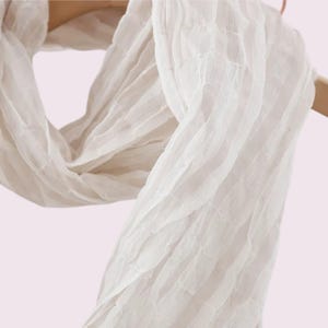 May include: A lightweight white scarf featuring a distinct crinkled texture and vertical striped pattern. The fabric is semi-sheer and draped, showcasing a series of horizontal ridges that create a pleated effect throughout the entire length of the accessory.
