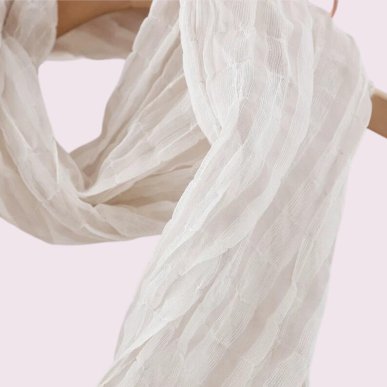 May include: A lightweight white scarf featuring a distinct crinkled texture and vertical striped pattern. The fabric is semi-sheer and draped, showcasing a series of horizontal ridges that create a pleated effect throughout the entire length of the accessory.