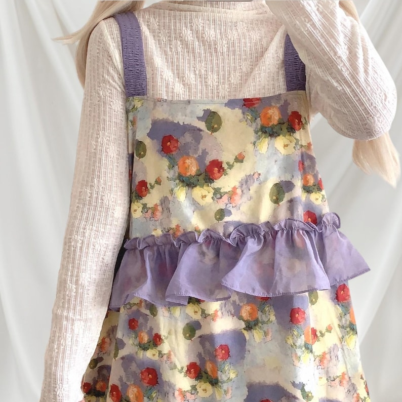 May include: A floral print dress with a light purple ruffle and straps. The dress features a watercolor-style pattern with red, orange, and yellow flowers on a light blue and yellow background. The straps are a solid light purple color.