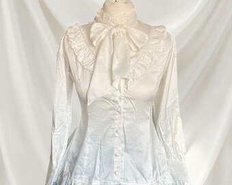May include: A long-sleeved white blouse featuring a high ruffled collar, a large bow tie at the neckline, and vertical ruffles extending from the shoulders to the chest. The garment includes a button-down front and a ruffled hemline at the waist.