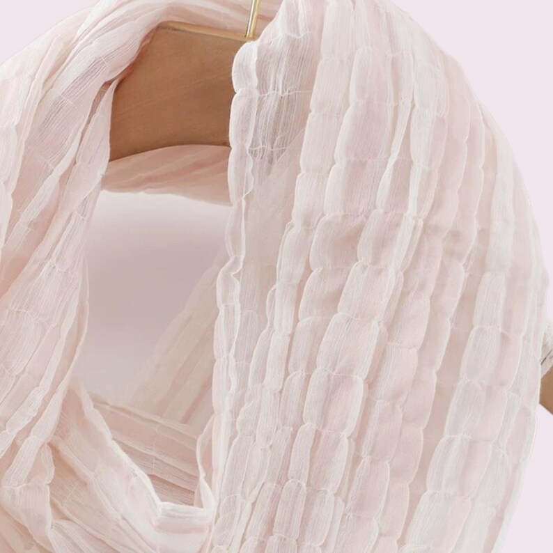 May include: A light pink infinity scarf made of a sheer, lightweight fabric with a distinct crinkled, pleated texture. The scarf features vertical rows of gathered material that create a soft, voluminous look and a consistent, repeating pattern.