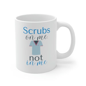Puede incluir: Taza de cerámica blanca con el texto "Scrubs on me, not in me" en fuente azul y negra. Un gráfico de uniforme médico azul claro está centrado debajo de las palabras "on me". La taza tiene un asa curva y es adecuada para bebidas frías o calientes.
