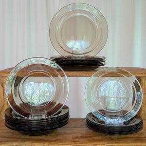 May include: A stack of 12 clear glass dinner plates with a brown rim. The plates are stacked on a wooden surface.