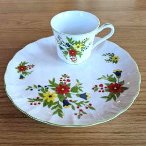 May include: White teacup and saucer set with a floral design. The cup and saucer have a scalloped edge with a green trim. The floral pattern includes red, yellow, and blue flowers with green leaves.