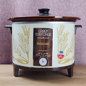 May include: A cream-colored Crock Watcher by Hamilton Beach 6-quart slow cooker with a brown lid and base. The exterior features yellow botanical illustrations of thyme and bay leaf, a wood-grain control panel, and a red Buy American sticker.