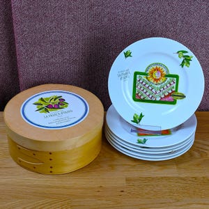 May include: A set of six white porcelain plates featuring olive branch illustrations and a central graphic with a sun and olive grove, labeled La Frasca d'Olivo. The plates are stacked beside a round wooden storage box with a matching olive label.