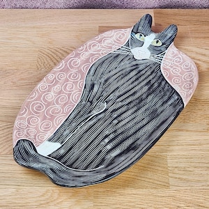 May include: An oval ceramic tray featuring a black and white cat illustration with green eyes. The cat is centered on a light pink background decorated with white spiral patterns. The tray has a smooth finish and a distinct cat-shaped silhouette.