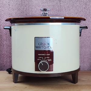 May include: A vintage Sears Kenmore Crock Watcher slow cooker with a cream-colored exterior, a dark brown base, and matching side handles. It features a glass lid with a clear knob and a control panel with settings for off, low, high, and auto shift.