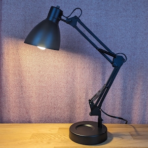Puede incluir: Una lámpara de escritorio negra ajustable con una base redonda y una pantalla cónica. La lámpara tiene un brazo flexible y está colocada sobre una superficie de madera, proyectando una luz cálida.