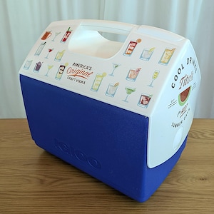 Puede incluir: Una nevera portátil Igloo azul y blanca con asa blanca. La nevera presenta ilustraciones de cócteles y el texto "America's Original Craft Vodka" y "Cool Drinks Tito's Summer Days".