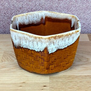 Puede incluir: Una maceta de cerámica marrón con un diseño de tejido de cesta. El borde superior tiene un esmalte blanco texturizado. La maceta tiene forma cuadrada con esquinas redondeadas. La maceta está sobre una superficie de madera.