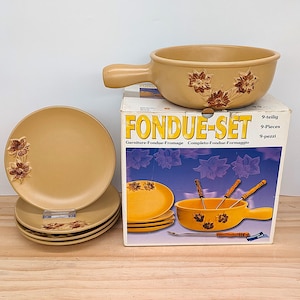 May include: A tan ceramic fondue set with floral accents. The set includes a fondue pot with a handle, plates, and forks. The box reads "FONDUE-SET" and "9-Pieces".