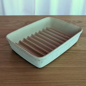 May include: Rectangular, off-white ceramic dish with a series of parallel ridges. The dish has a slightly distressed appearance, suggesting it is vintage or handmade. The ridges are a light brown color. The dish is likely used for draining items.