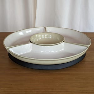 May include: A white and beige ceramic serving dish set. The set includes a central round bowl surrounded by three crescent-shaped compartments. The base is a dark grey colour. The set is on a wooden surface.