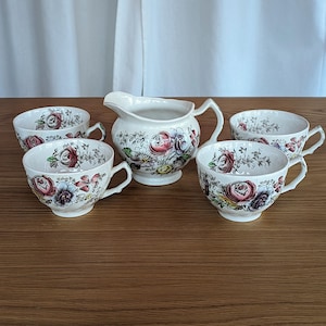 May include: A cream-colored tea set featuring a floral pattern with red, pink, and purple roses. The set includes four teacups and a creamer pitcher. The cups have curved handles, and the pitcher has a spout and handle.