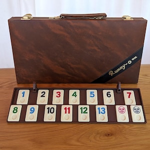 May include: A brown Rummy-O game set with a matching carrying case. The game board displays numbered tiles from 1 to 13, with two joker tiles. The case has a handle and gold-colored hardware.