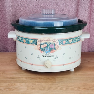 Puede incluir: Olla de cocción lenta Rival de color crema con tapa transparente y borde verde oscuro. La olla de cocción lenta tiene una banda decorativa con un diseño floral y de frutas y el texto "A Garden of Good Things".