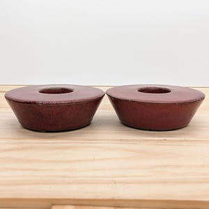 May include: Two maroon-colored ceramic candle holders. Each has a circular design with a hole in the center. The candle holders are on a light-colored wooden surface.
