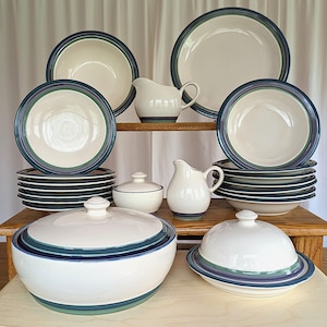 May include: A set of white ceramic dinnerware with blue and green stripes. The set includes a large serving bowl with a lid, a butter dish with a lid, two creamers, a sugar bowl with a lid, and a stack of dinner plates.