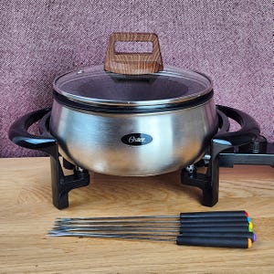 May include: A stainless steel Oster electric fondue pot with a glass lid featuring a wood-grain handle. The pot has black side handles and a temperature control dial. Six metal fondue forks with black handles and colored tips are arranged in front.