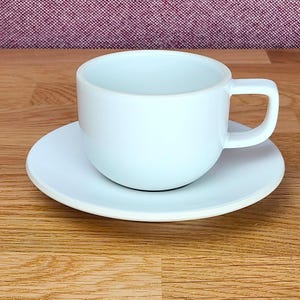 Può includere: Una tazza da tè in ceramica bianca con base arrotondata e piattino abbinato. La tazza ha un manico e poggia sul piattino. Il set è su una superficie di legno.