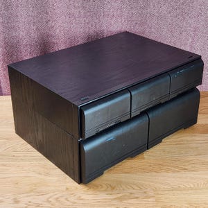 Può includere: Un'unità di archiviazione da scrivania in legno nero con una superficie superiore piatta e quattro cassetti disposti su due file. L'unità ha una forma rettangolare con una venatura del legno visibile sui pannelli laterali e frontali.