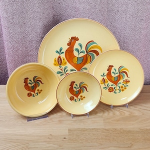 May include: A set of four ceramic dishes in a cream color with a speckled finish. The collection includes two plates and two bowls, each featuring a central illustration of a rooster in orange, yellow, and teal tones, surrounded by floral motifs.