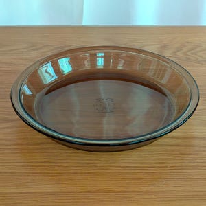 May include: A clear, oval-shaped pie dish made of brown-tinted glass. The dish has a slightly raised rim and a smooth, flat bottom. The dish is sitting on a wooden surface.