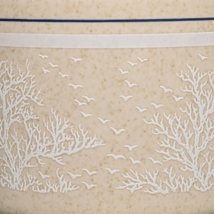 May include: A close-up of a ceramic mug with a beige background and a white design of trees and birds. The mug has a blue stripe around the top.