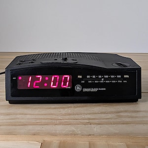 May include: Black GE FM/AM clock radio with a digital display showing 12:00 in pink. The radio has a tuning dial and buttons on top. The text "FM/AM Clock Radio Battery Backup" is visible.