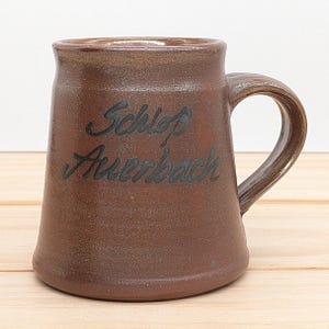 May include: A brown ceramic mug with a black handle. The mug has the words "Schloß Auerbach" written on it in black.
