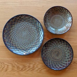 Op de afbeelding: Drie keramische borden met een getextureerde, iriserende afwerking. De borden zijn in tinten blauw, groen en bruin, met een spiraalpatroon dat vanuit het midden uitstraalt. De borden zijn op een houten oppervlak geplaatst.