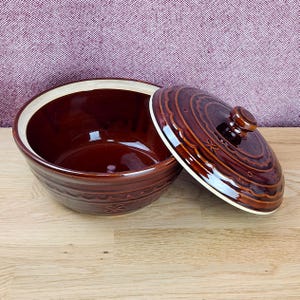 May include: A round, deep ceramic bowl with a glossy dark brown glaze and a matching lid. The exterior features a decorative embossed wavy pattern and small flower motifs. The interior of the bowl and the underside of the lid are cream colored.