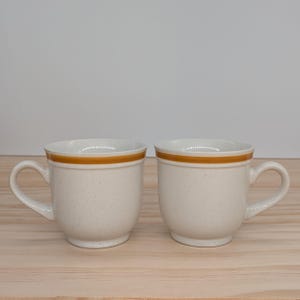 May include: Two white ceramic mugs with a brown band around the top. The mugs have a speckled finish.