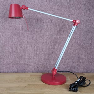 May include: A red desk lamp with a circular base and adjustable arm. The lamp features a red lampshade and white and red support arms. The lamp is on a wooden surface with a purple background.