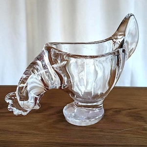 May include: A clear glass egg cup shaped like a rooster. The cup has a curved neck and a round base.