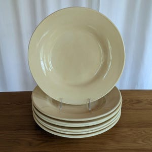 May include: A stack of six cream-colored ceramic plates. One plate is upright, and the others are stacked below. The plates have a simple, classic design with a slightly raised rim. The plates are on a wooden surface.