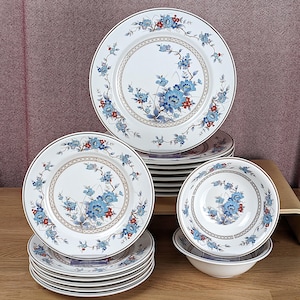 Puede incluir: Un juego de vajilla de porcelana blanca con un diseño floral en azul y rojo. El juego incluye platos y cuencos, con un borde marrón. El estampado floral está centrado en cada pieza.