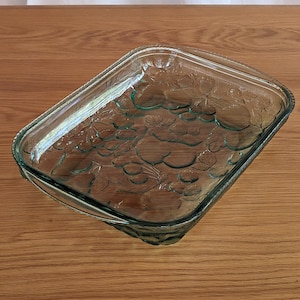 Op de afbeelding: Een heldere glazen ovenschaal met een rechthoekige vorm en afgeronde hoeken. De binnenkant heeft een verhoogd druiven- en bladerontwerp. De schaal heeft een getextureerde rand en is geschikt voor gebruik in de oven.