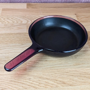 Può includere: Una piccola padella nera con un lungo manico bordeaux. Il bordo della padella è anch'esso bordeaux. La padella è appoggiata su una superficie di legno chiaro. La padella è progettata per cucinare.