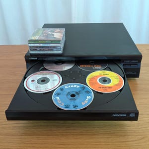 May include: A black Yamaha CD player with a disc tray holding five CDs. The tray is open, displaying the CDs with various album art. A stack of CDs rests on top of the player.