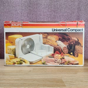 May include: A white Krups Universal Compact food slicer shown on its original cardboard packaging. The box features a graphic illustration of the appliance alongside various cheeses, breads, and cured meats arranged on a yellow surface.