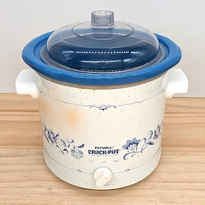 Puede incluir: Una olla de cocción lenta de gres Rival Crock-Pot con tapa transparente y detalles azules. La olla blanca presenta un diseño floral en azul y un mando con ajustes de apagado, bajo y alto. Asas blancas a cada lado.