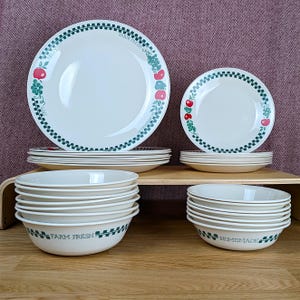 Puede incluir: Una colección de vajilla de cerámica blanca con un borde a cuadros verde y negro. Los platos y cuencos presentan diseños de frutas y flores. Algunos cuencos están etiquetados como "Farm Fresh" y "Homemade".