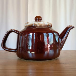 Puede incluir: Una tetera de cerámica marrón con un borde blanco y un pomo marrón en la tapa. La tetera tiene un acabado brillante y está sobre una superficie de madera.